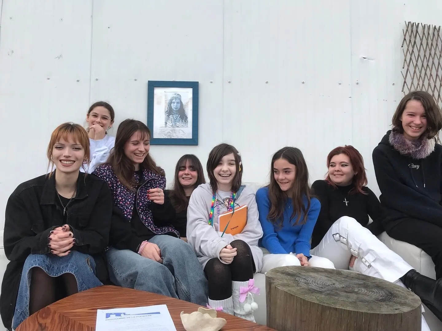 Un groupe de huit jeunes souriants assis sur un banc devant le mur blanc du lieu, un portrait au crayon encadré derrière eux