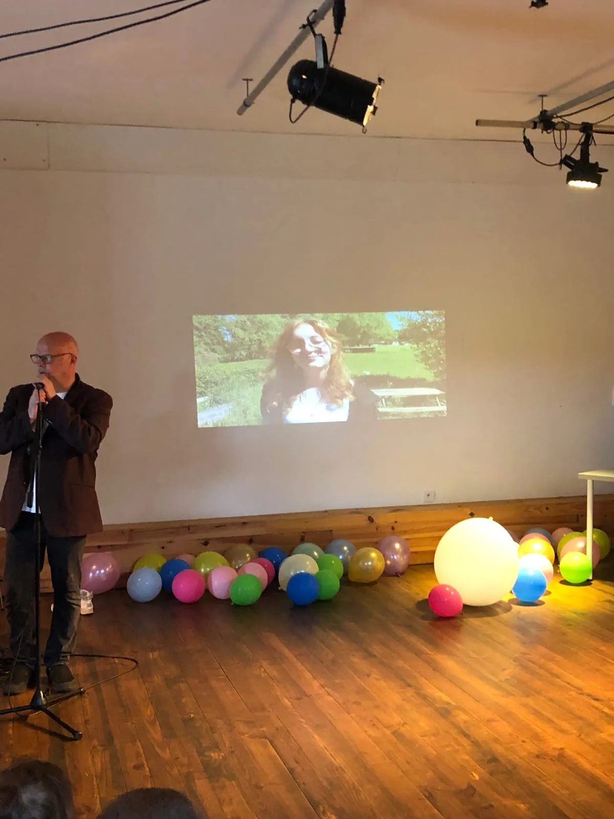 Un homme au micro sur scène, une vidéo projetée sur le mur derrière lui et des ballons colorés éparpillés sur le parquet