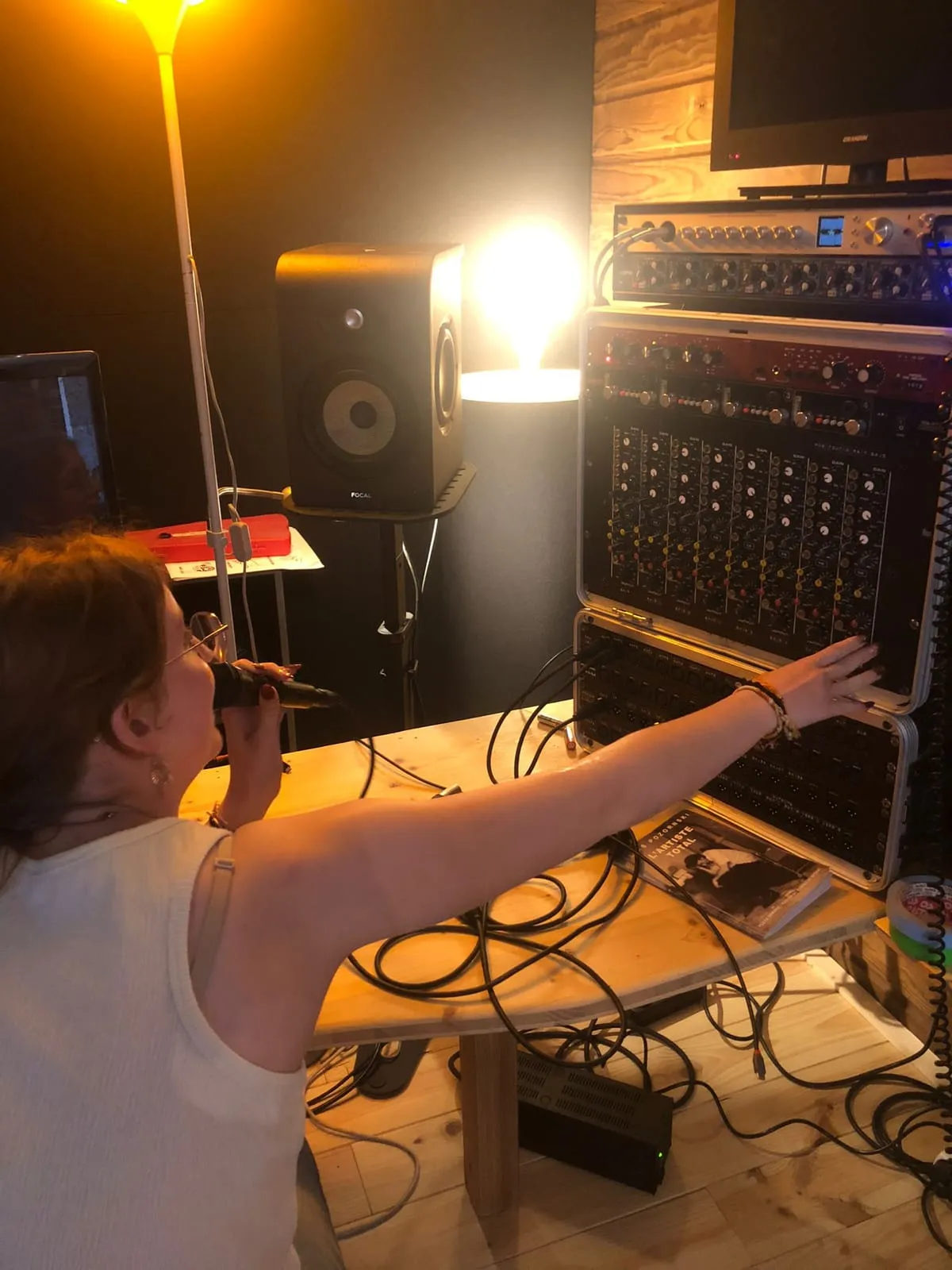 Une jeune femme ajustant un micro devant une enceinte de monitoring, une table de mixage et un rack d'effets derrière elle
